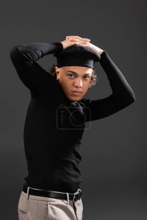 fashionable african american man in black beret and turtleneck posing with hands on head isolated on dark grey