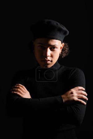 hombre afroamericano con estilo en boina y cuello alto de pie con los brazos cruzados y mirando a la cámara aislada en negro