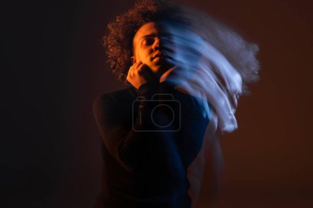 double exposure of injured african american man with hands near bleeding face standing with closed eyes on dark background with red and blue light
