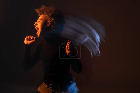 Foto de Larga exposición de hombre afroamericano enojado y estresado con trastorno bipolar gritando sobre fondo negro con luz naranja y azul - Imagen libre de derechos