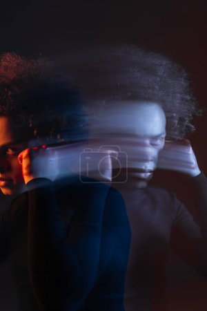 Foto de Doble exposición del hombre afroamericano abusado con la mano cerca de la cara lesionada mirando a la cámara en la oscuridad con luz roja y azul - Imagen libre de derechos