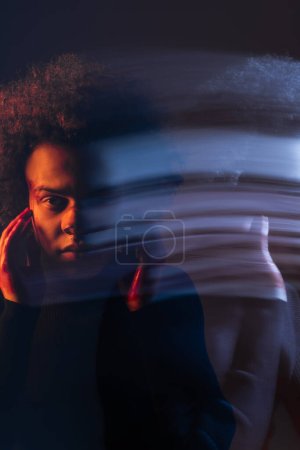 Doppelbelichtung eines misshandelten afrikanisch-amerikanischen Mannes mit bipolarer Störung, der verletztes blutendes Gesicht auf dunklem Hintergrund mit orangefarbenem und blauem Licht berührt
