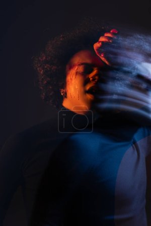 Doppelbelichtung des verwundeten afrikanisch-amerikanischen Mannes, der mit der Hand in der Nähe des blutenden Gesichts auf dunklem Hintergrund mit rotem und blauem Licht steht