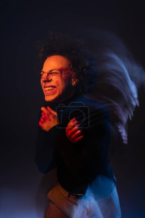 motion blur of wounded african american man with bipolar disorder and bleeding face grimacing on dark with orange and blue light
