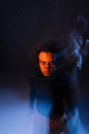 wounded african american man with bipolar disorder and bloody face looking at camera on dark background with orange and blue light