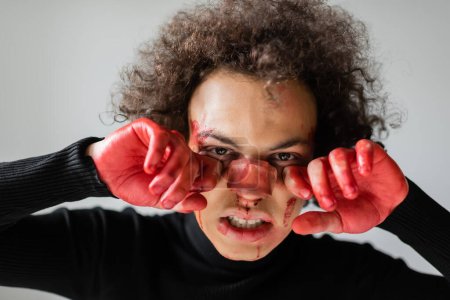 hombre afroamericano herido sosteniendo manos ensangrentadas cerca de la cara con la nariz rota aislada en gris