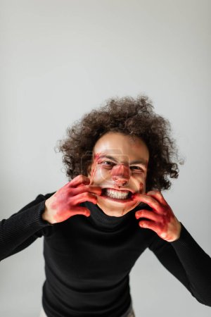 Foto de Wounded and angry african american man with bleeding face and broken nose grimacing at camera isolated on grey - Imagen libre de derechos