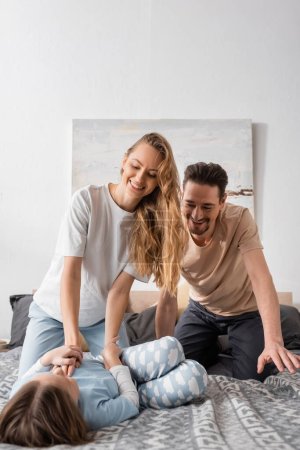 happy woman tickling daughter near smiling husband on bed 