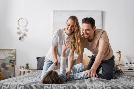 happy woman tickling daughter near cheerful husband on bed 