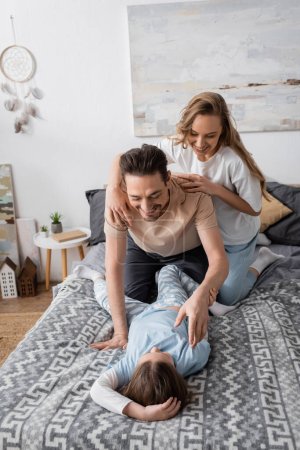 happy man tickling daughter near cheerful wife on bed 