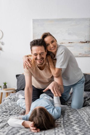 cheerful woman hugging happy husband laughing while having fun with child at home 