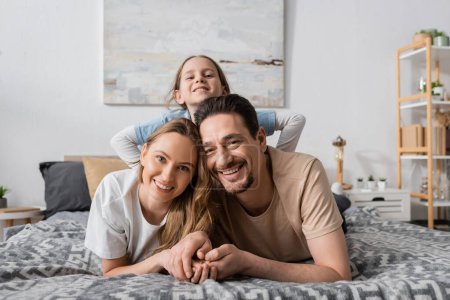 portrait of happy child behind positive parents looking at camera while resting in bedroom 