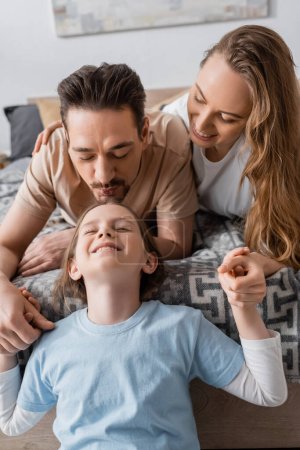 bearded man kissing forehead of cheerful daughter near wife 