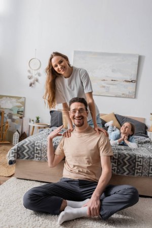 Photo for Woman leaning on shoulders of happy husband sitting on carpet near daughter on bed - Royalty Free Image