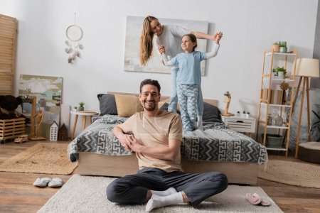 Foto de Cheerful bearded man sitting on carpet near wife and daughter standing on bed on blurred background - Imagen libre de derechos