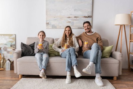 Photo for Full length of cheerful family holding glasses of orange juice and sitting on sofa - Royalty Free Image