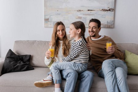 cheerful daughter sitting on laps of mother near positive father holding glass of orange juice 