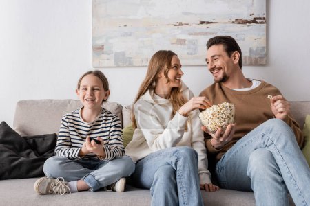 Photo for Cheerful parents eating popcorn near happy daughter with remote controller - Royalty Free Image