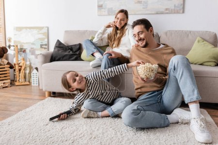 Photo for Happy kid with remote controller reaching popcorn near father and mother on blurred background - Royalty Free Image