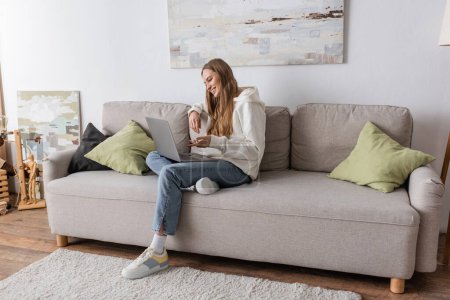 Photo for Full length of positive woman in casual clothes having video call on laptop in living room - Royalty Free Image