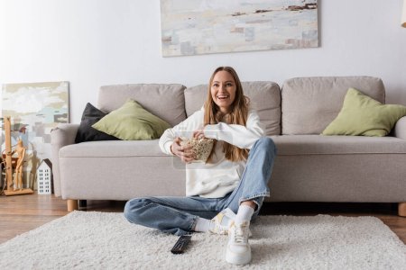 full length of joyful woman holding bowl with popcorn while watching movie in living room