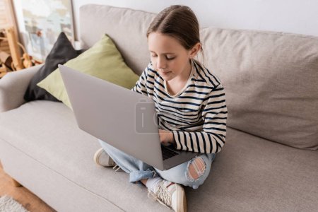 Téléchargez les photos : High angle view of preteen girl in casual clothes using laptop while sitting on couch - en image libre de droit