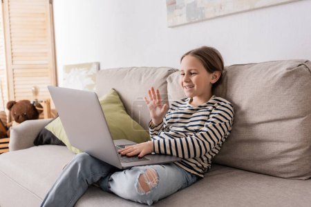preteen girl in casual clothes having video call on laptop while sitting on couch 