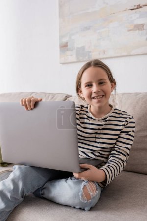 Photo for Joyful preteen girl in casual clothes looking at camera and holding laptop while sitting on couch - Royalty Free Image