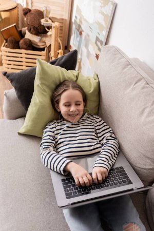 high angle view of smiling preteen girl in casual clothes using laptop while lying on couch 