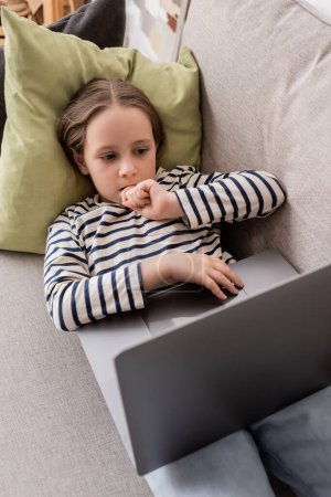 high angle view of focused preteen girl in casual clothes using laptop while lying on couch 