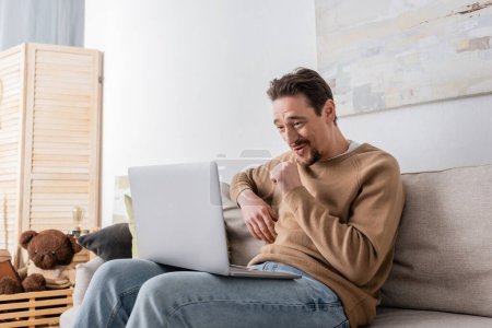Téléchargez les photos : Bearded man watching movie on laptop while sitting on sofa in living room - en image libre de droit
