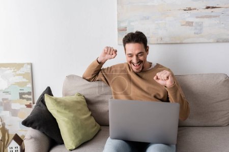 positive man watching championship on laptop while cheering and sitting on sofa in living room 