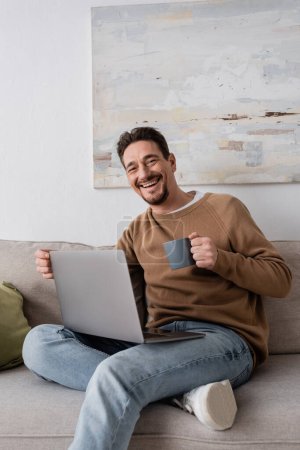 joyful man using laptop while holding cup and sitting on sofa in living room 