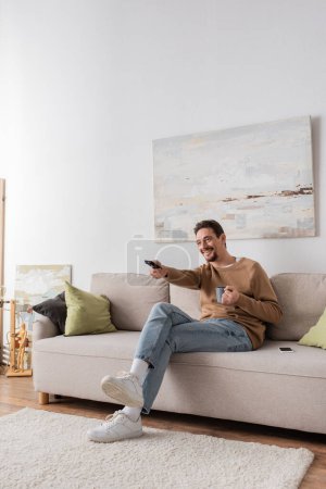 full length of cheerful man holding remote controller and cup of coffee in living room 