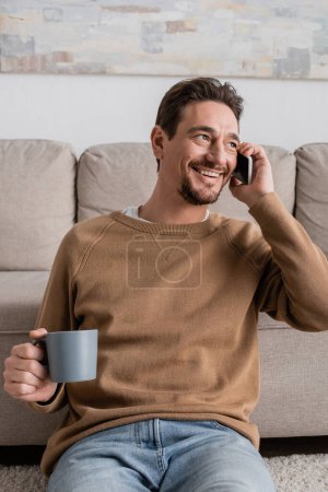 smiling man in beige jumper talking on smartphone while sitting with cup of coffee at home 