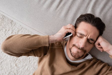 top view of exhausted man in beige jumper holding mobile phone and leaning on sofa 