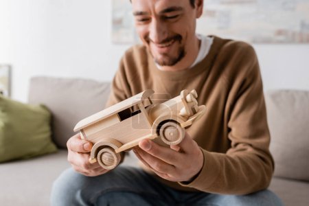 Photo for Bearded man smiling while looking at wooden car toy in living room - Royalty Free Image