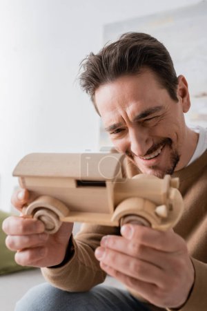 Photo for Cheerful man smiling while looking at wooden car toy in living room - Royalty Free Image