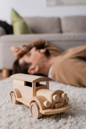 Photo for Wooden handmade car toy near blurred man lying on carpet in living room - Royalty Free Image