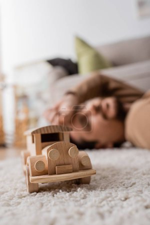 Photo for Wooden toy car near blurred man lying on carpet in living room - Royalty Free Image