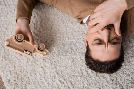 Photo for Top view of sad man covering mouth with hand lying on carpet near wooden toy vehicle in living room - Royalty Free Image
