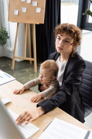 businesswoman sitting with little daughter at workplace and using computer near documents on desk