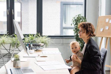 happy businesswoman sitting at workplace with little daughter and looking at computer monitor