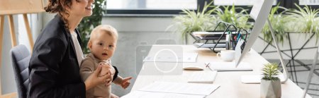 toddler child looking at camera near smiling mother sitting at workplace near documents and computer monitor, banner