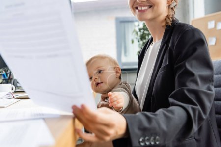 joyful businesswoman holding blurred document near little child with outstretched hand in office