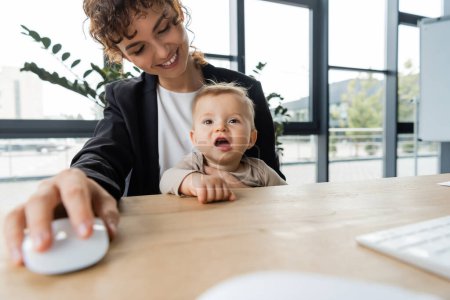 cheerful businesswoman sitting at workplace with little child and holding blurred computer mouse