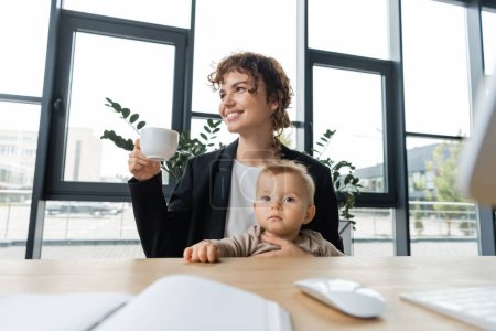 happy businesswoman sitting with little daughter and coffee cup near blurred computer mouse and notebook on work desk