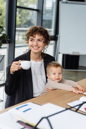 smiling businesswoman looking away while sitting with toddler child and coffee cup at workplace in office