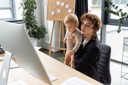 businesswoman in black blazer holding baby and looking at computer monitor in office