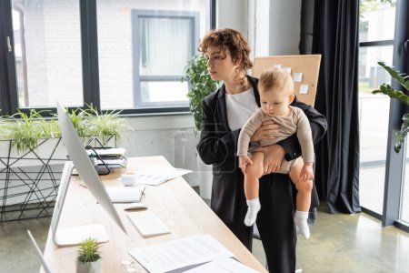 businesswoman in black suit looking at computer monitor while standing with little daughter at work desk in office